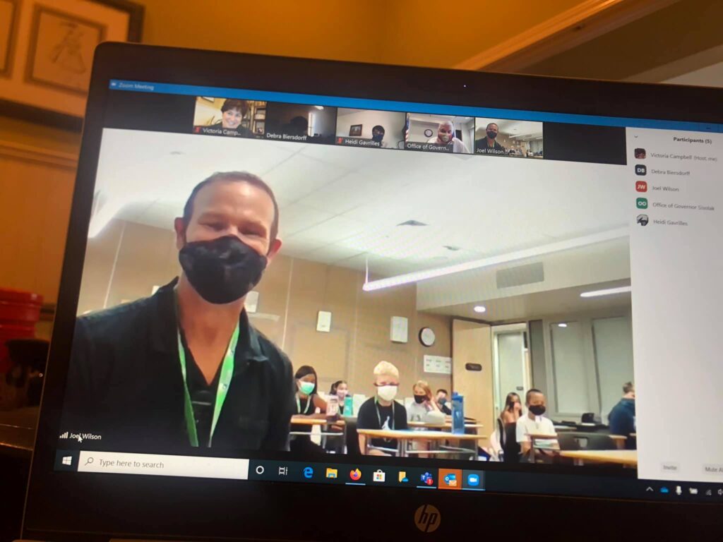 Teacher wearing a mask can be seen via Zoom on computer screen. Behind him is a class of students also wearing masks.