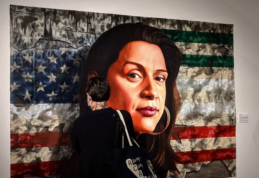 A woman looks closely at a painting of a woman's face in front of U.S. and Mexican flags.