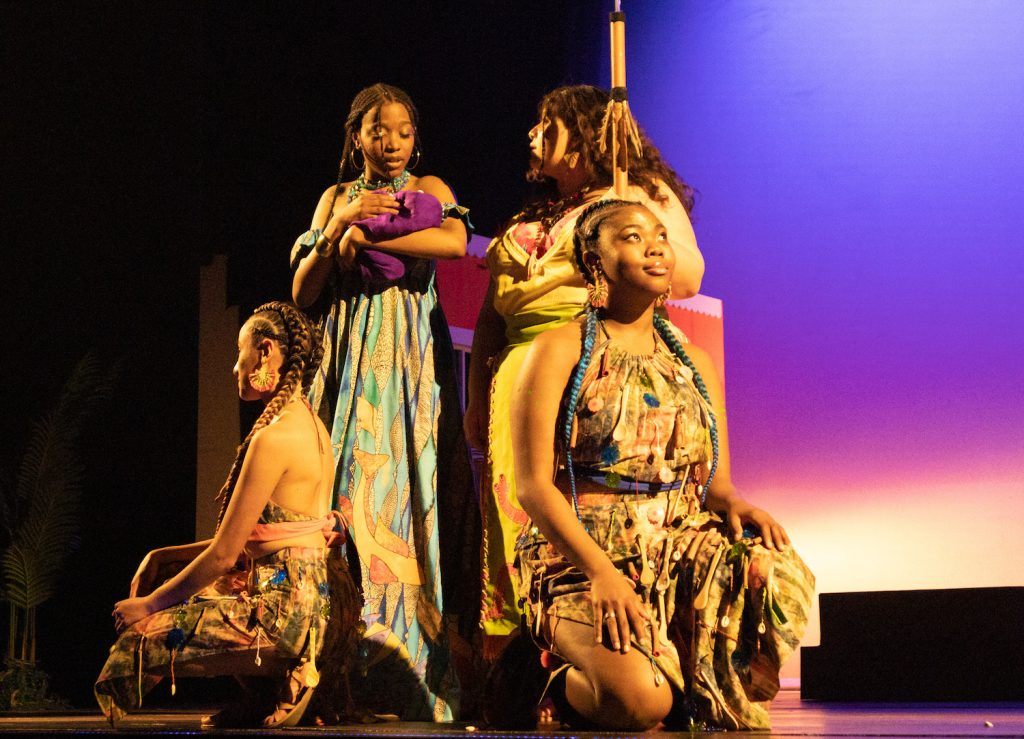 Four women on stage, two seat, one holds baby in blanket