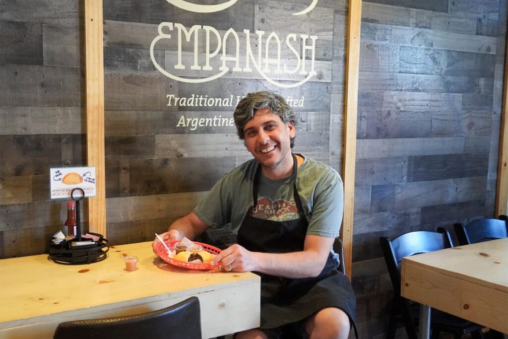 Man pictures sits holding empanada at a restauraunt, smiling