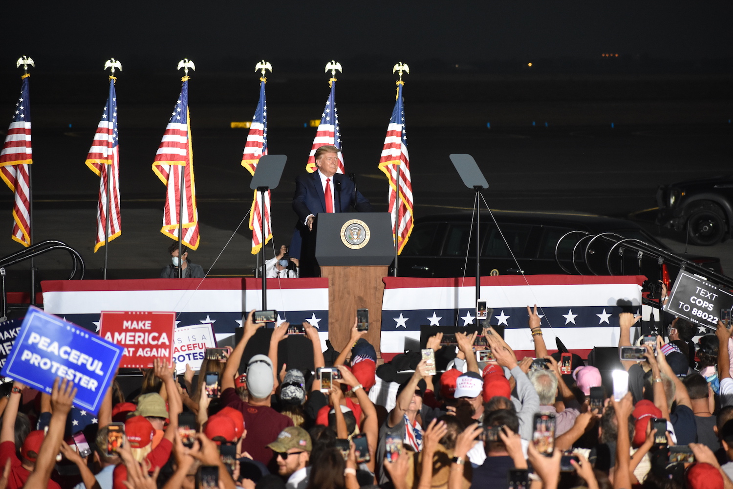 President Donald Trump stands behind a podium in front of four U.S. flags on a platform in front of attendees at the Minden-Tahoe Airport in Nevada.