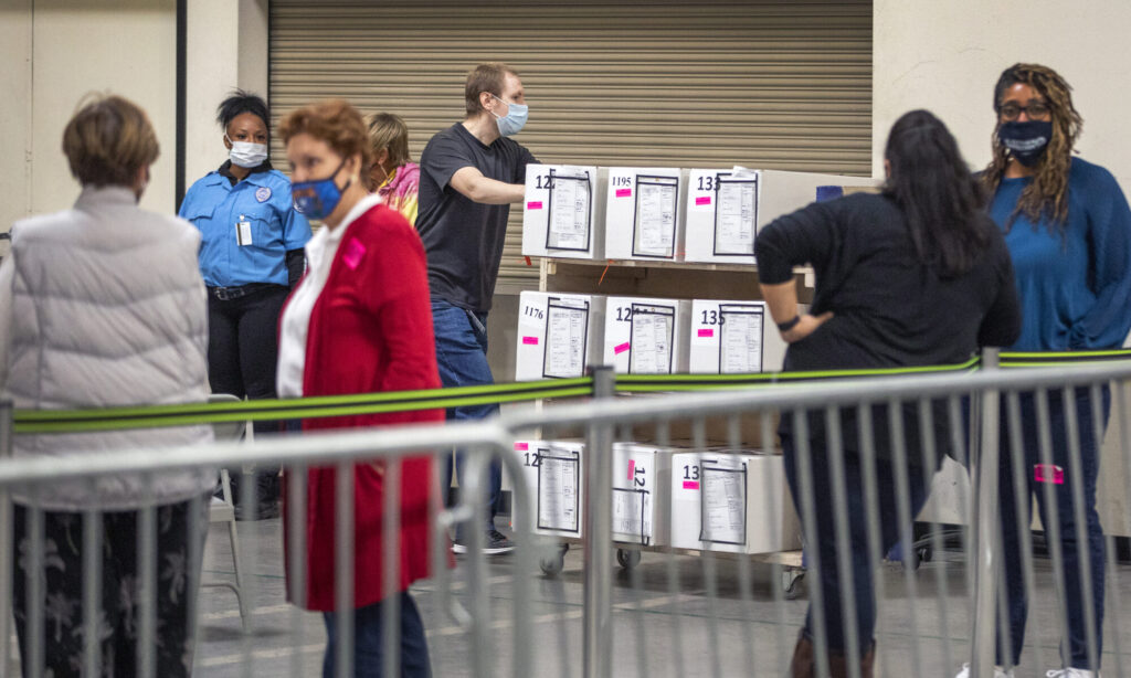 A masked man pushes a cart full of ballots pass observers