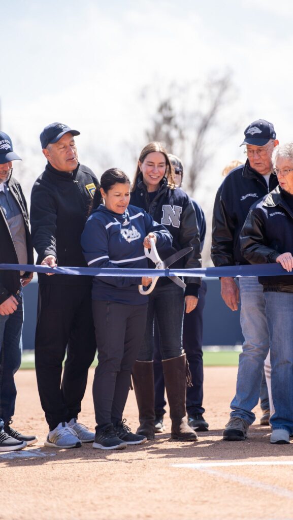 Photo of Linda Garza holding large scissors and cutting a ribbon at the UNR softball field. 